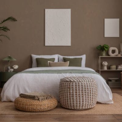 Aesthetic composition of stylish bedroom interior with mock up poster frame, cozy bed, braided pouf, rack,  gray armchair, vase with leaves, plaid and personal accessories. Home decor. Template.